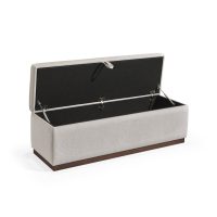 Banqueta retangular com topo almofadado, revestido em tecido bege, com uma base de madeira. Inclui um puxador em tecido na parte frontal para abrir o compartimento interno de arrumação, combinando funcionalidade e estilo.
