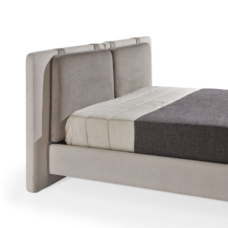 Cama moderna com cabeceira almofadada em tecido cinza-claro, composta por duas grandes almofadas anexadas. A estrutura da cama é estofada no mesmo tecido cinza-claro, com lençóis bege e cobertor cinza-escuro. A cama possui pés metálicos elegantes que a elevam ligeiramente do chão