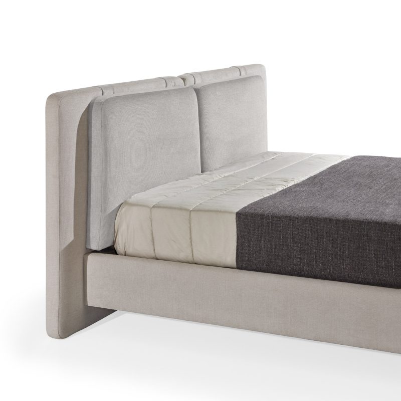 Cama moderna com cabeceira almofadada em tecido cinza-claro, composta por duas grandes almofadas anexadas. A estrutura da cama é estofada no mesmo tecido cinza-claro, com lençóis bege e cobertor cinza-escuro. A cama possui pés metálicos elegantes que a elevam ligeiramente do chão