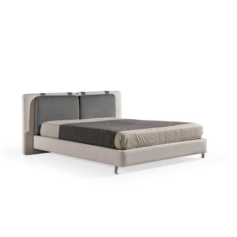 Cama moderna com cabeceira almofadada em tecido cinza-claro, composta por duas grandes almofadas anexadas. A estrutura da cama é estofada no mesmo tecido cinza-claro, com lençóis bege e cobertor cinza-escuro. A cama possui pés metálicos elegantes que a elevam ligeiramente do chão