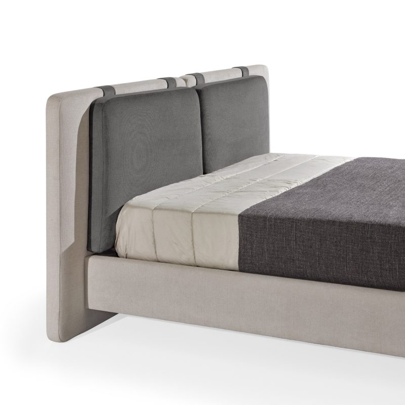 Cama moderna com cabeceira almofadada em tecido cinza-claro, composta por duas grandes almofadas anexadas. A estrutura da cama é estofada no mesmo tecido cinza-claro, com lençóis bege e cobertor cinza-escuro. A cama possui pés metálicos elegantes que a elevam ligeiramente do chão