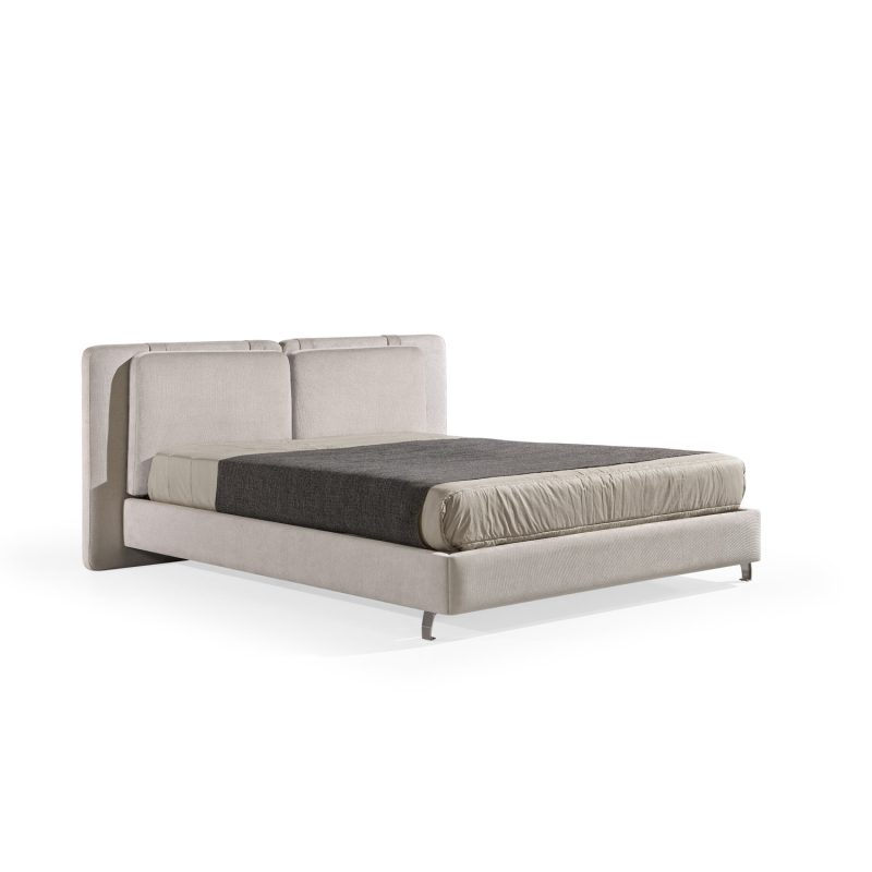 Cama moderna com cabeceira almofadada em tecido cinza-claro, composta por duas grandes almofadas anexadas. A estrutura da cama é estofada no mesmo tecido cinza-claro, com lençóis bege e cobertor cinza-escuro. A cama possui pés metálicos elegantes que a elevam ligeiramente do chão