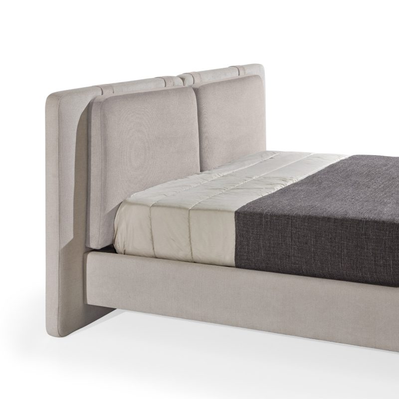 Cama moderna com cabeceira almofadada em tecido cinza-claro, composta por duas grandes almofadas anexadas. A estrutura da cama é estofada no mesmo tecido cinza-claro, com lençóis bege e cobertor cinza-escuro. A cama possui pés metálicos elegantes que a elevam ligeiramente do chão