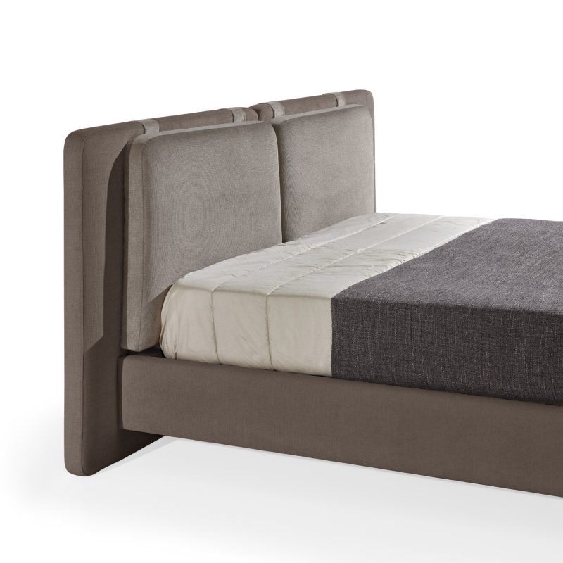 Cama moderna com cabeceira almofadada em tecido castanho, composta por duas grandes almofadas anexadas. A estrutura da cama é estofada no mesmo tecido castanho, com lençóis bege e cobertor cinza-escuro. A cama possui pés metálicos elegantes que a elevam ligeiramente do chão