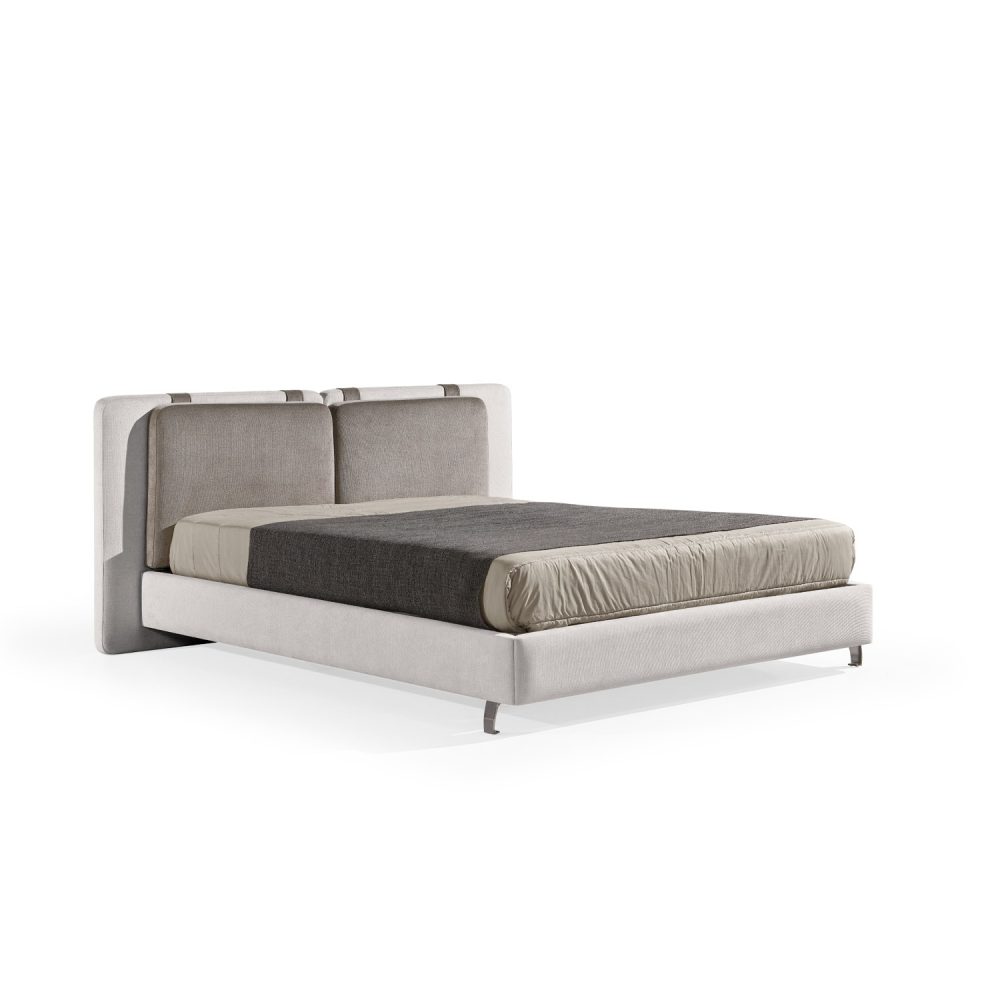 Cama moderna com cabeceira almofadada em tecido cinza-claro, composta por duas grandes almofadas anexadas. A estrutura da cama é estofada no mesmo tecido cinza-claro, com lençóis bege e cobertor cinza-escuro. A cama possui pés metálicos elegantes que a elevam ligeiramente do chão