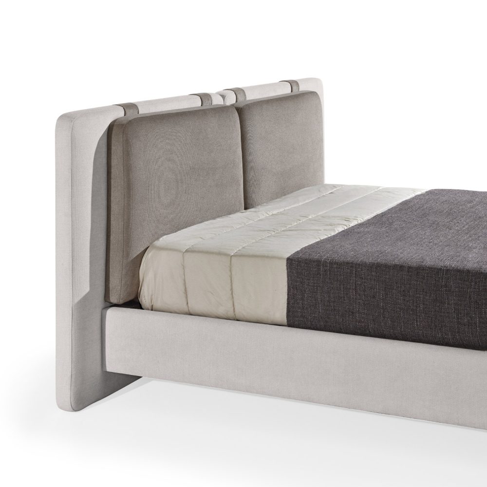 Cama moderna com cabeceira almofadada em tecido cinza-claro, composta por duas grandes almofadas anexadas. A estrutura da cama é estofada no mesmo tecido cinza-claro, com lençóis bege e cobertor cinza-escuro. A cama possui pés metálicos elegantes que a elevam ligeiramente do chão