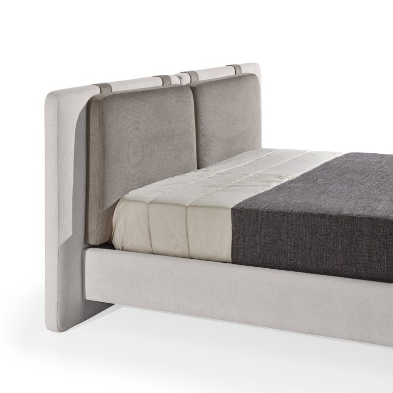 Cama moderna com cabeceira almofadada em tecido cinza-claro, composta por duas grandes almofadas anexadas. A estrutura da cama é estofada no mesmo tecido cinza-claro, com lençóis bege e cobertor cinza-escuro. A cama possui pés metálicos elegantes que a elevam ligeiramente do chão