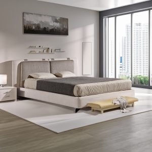 Quarto moderno com uma cama estofada de tom claro e cabeceira alta, adornada com dois travesseiros e colchas em tons de bege e cinzento. À esquerda, há uma mesa de cabeceira branca com um candeeiro, enquanto acima da cama está uma obra de arte abstrata em tons de cinza, branco e preto. Abaixo da obra, pequenas prateleiras exibem livros e peças decorativas. Em frente à cama, encontra-se um banco baixo com um tecido decorativo por cima. À direita, uma grande janela oferece vista para edifícios altos e vegetação.
