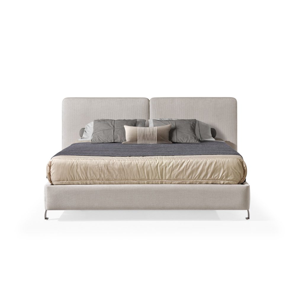 Cama moderna com cabeceira acolchoada de grandes dimensões, estrutura baixa em branco, colchão bege e roupa de cama com cobertor cinza-escuro e almofadas em tons de bege e cinza. A cama tem pés metálicos pequenos que conferem um design elegante e contemporâneo.