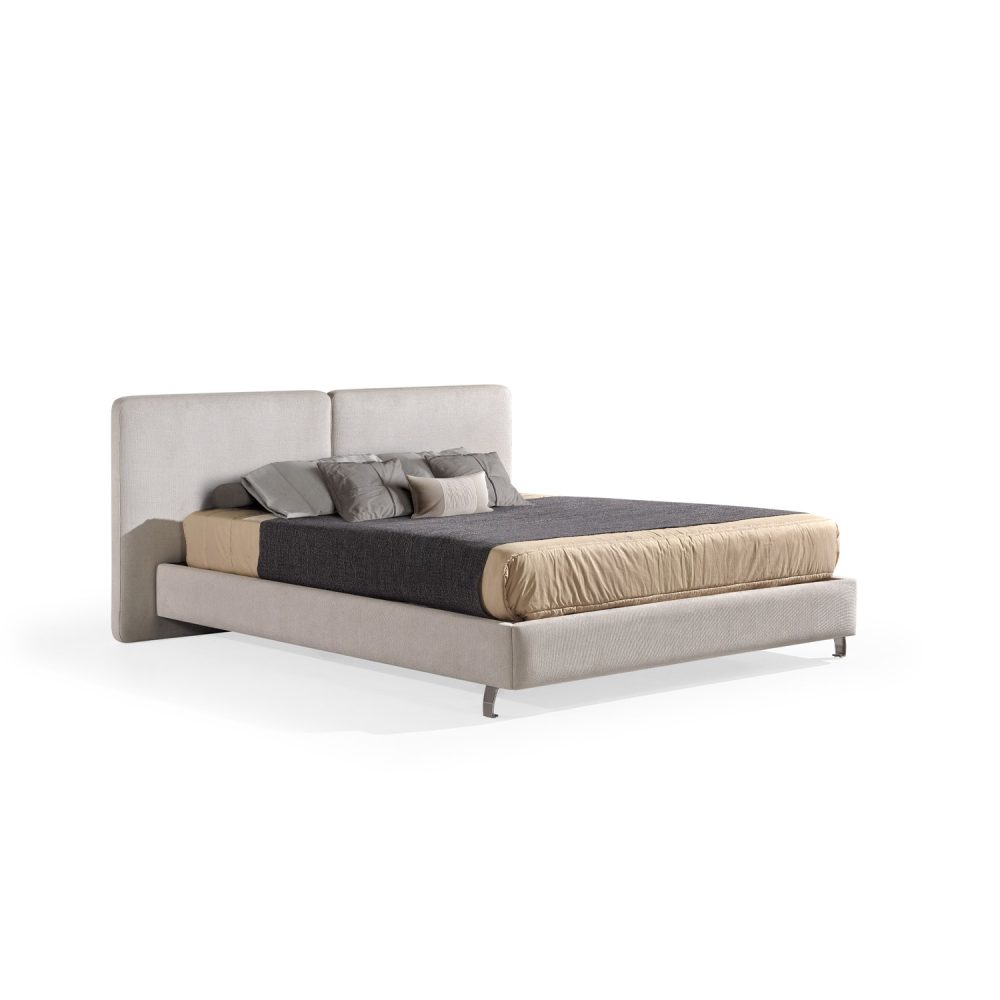 Cama moderna com cabeceira acolchoada de grandes dimensões, estrutura baixa em branco, colchão bege e roupa de cama com cobertor cinza-escuro e almofadas em tons de bege e cinza. A cama tem pés metálicos pequenos que conferem um design elegante e contemporâneo.