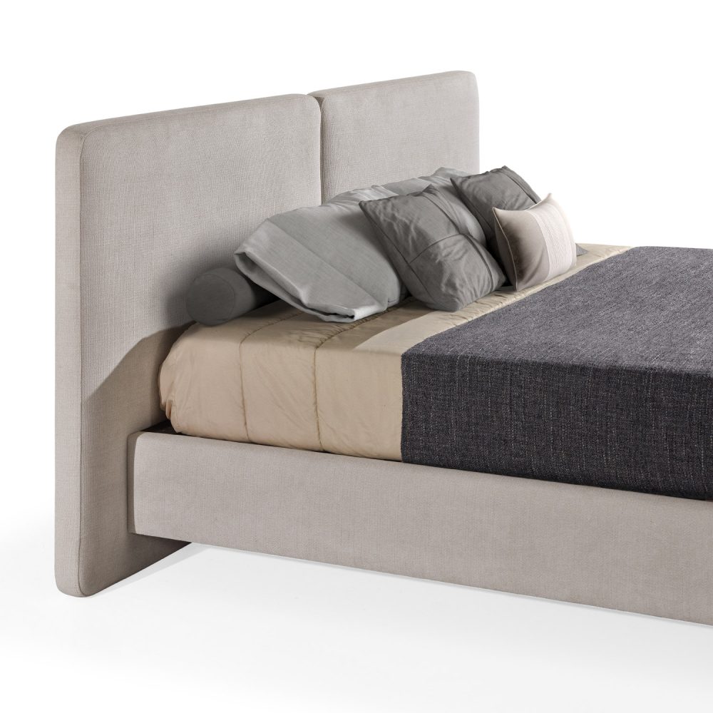 Cama moderna com cabeceira acolchoada de grandes dimensões, estrutura baixa em branco, colchão bege e roupa de cama com cobertor cinza-escuro e almofadas em tons de bege e cinza. A cama tem pés metálicos pequenos que conferem um design elegante e contemporâneo.
