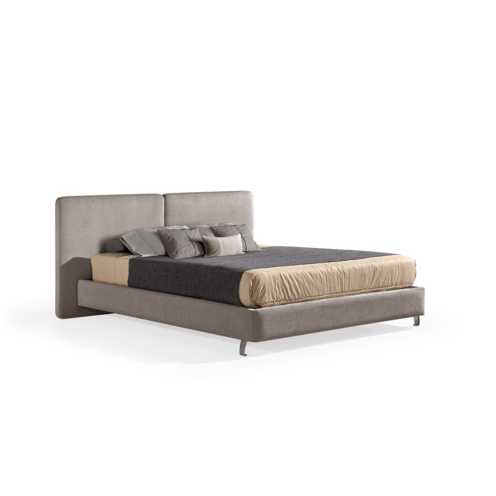 Cama moderna com cabeceira acolchoada de grandes dimensões, estrutura baixa castanho claro, colchão bege e roupa de cama com cobertor cinza-escuro e almofadas em tons de bege e cinza. A cama tem pés metálicos pequenos que conferem um design elegante e contemporâneo.