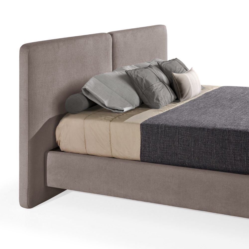 Cama moderna com cabeceira acolchoada de grandes dimensões, estrutura baixa castanho, colchão bege e roupa de cama com cobertor cinza-escuro e almofadas em tons de bege e cinza. A cama tem pés metálicos pequenos que conferem um design elegante e contemporâneo.