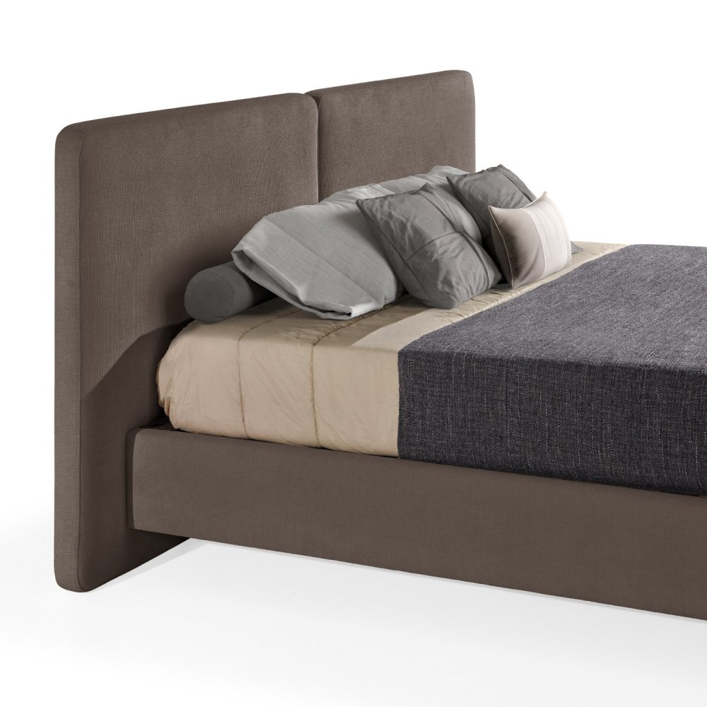 Cama moderna com cabeceira acolchoada de grandes dimensões, estrutura baixa em bege, colchão bege e roupa de cama com cobertor cinza-escuro e almofadas em tons de bege e cinza. A cama tem pés metálicos pequenos que conferem um design elegante e contemporâneo.