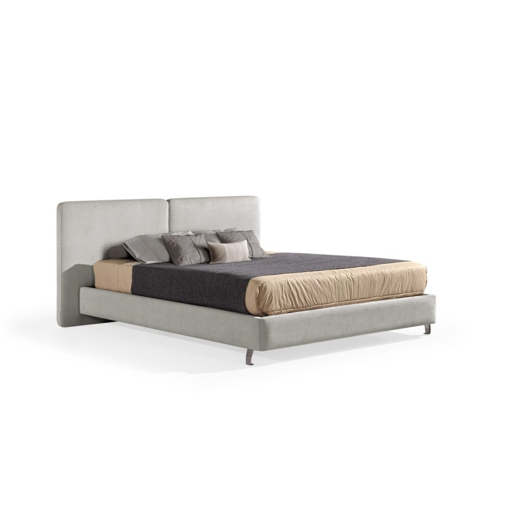 Cama moderna com cabeceira acolchoada de grandes dimensões, estrutura baixa bege, colchão bege e roupa de cama com cobertor cinza-escuro e almofadas em tons de bege e cinza. A cama tem pés metálicos pequenos que conferem um design elegante e contemporâneo.