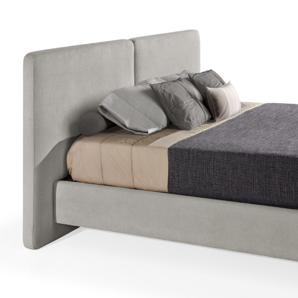Cama moderna com cabeceira acolchoada de grandes dimensões, estrutura baixa bege, colchão bege e roupa de cama com cobertor cinza-escuro e almofadas em tons de bege e cinza. A cama tem pés metálicos pequenos que conferem um design elegante e contemporâneo.
