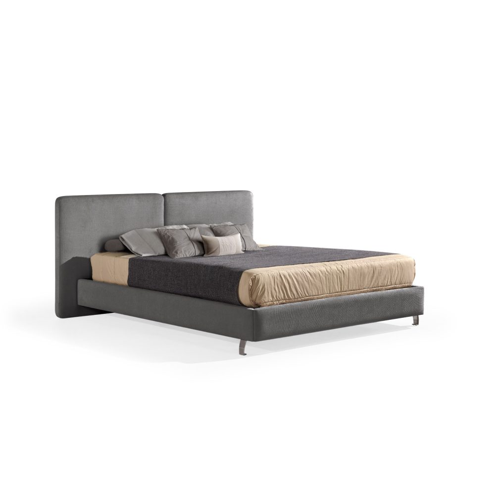 Cama moderna com cabeceira acolchoada de grandes dimensões, estrutura baixa em cinza-escuro, colchão bege e roupa de cama com cobertor cinza-escuro e almofadas em tons de bege e cinza. A cama tem pés metálicos pequenos que conferem um design elegante e contemporâneo.