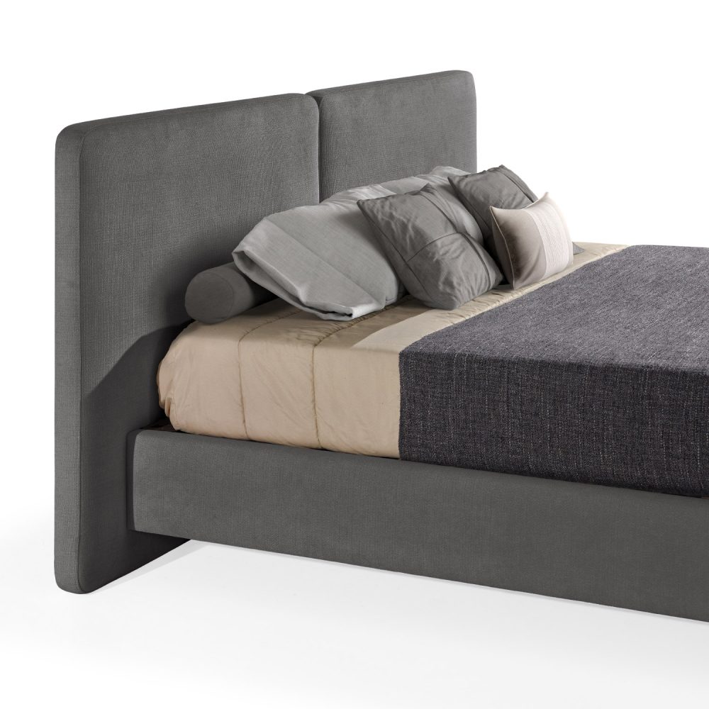 Cama moderna com cabeceira acolchoada de grandes dimensões, estrutura baixa em cinza-escuro, colchão bege e roupa de cama com cobertor cinza-escuro e almofadas em tons de bege e cinza. A cama tem pés metálicos pequenos que conferem um design elegante e contemporâneo.