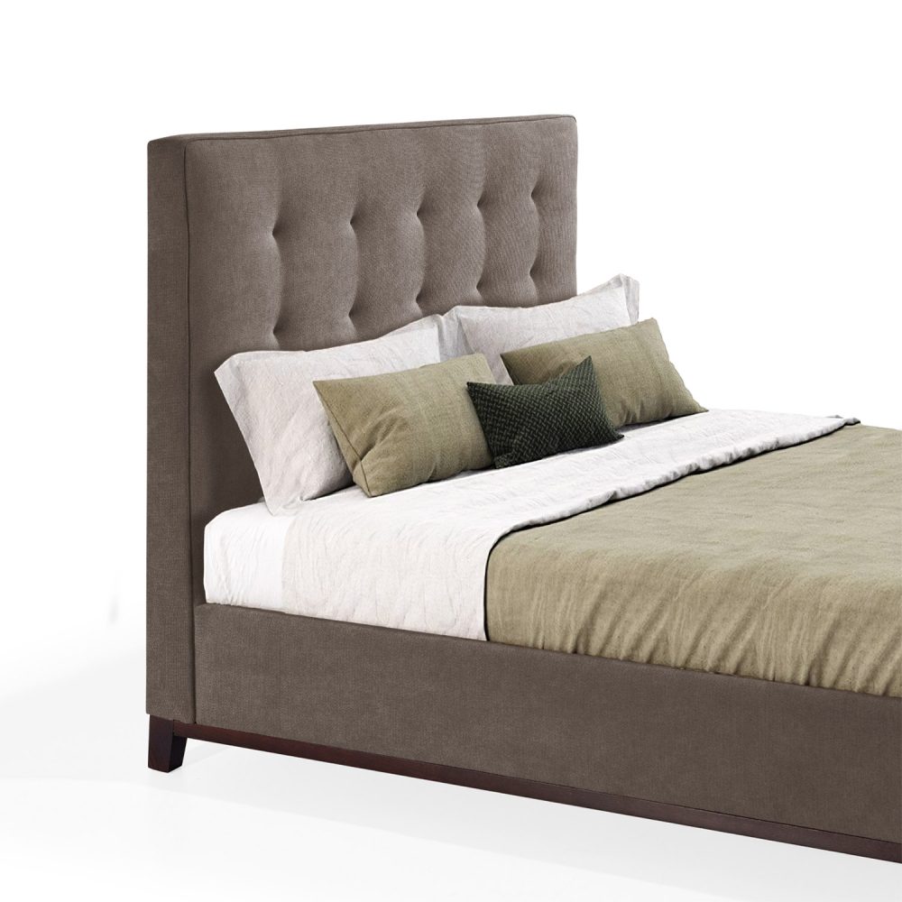 Cama moderna com cabeceira acolchoada e estofada em tecido castanho, com lençóis brancos, uma manta verde e várias almofadas em tons de verde e branco. A estrutura da cama é revestida com tecido castanho e possui pés de madeira escura.