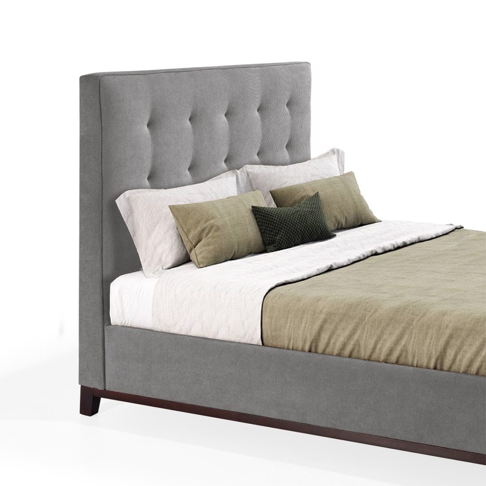 Cama moderna com cabeceira acolchoada e estofada em tecido cinza, com lençóis brancos, uma manta verde e várias almofadas em tons de verde e branco. A estrutura da cama é revestida com tecido cinza e possui pés de madeira escura.